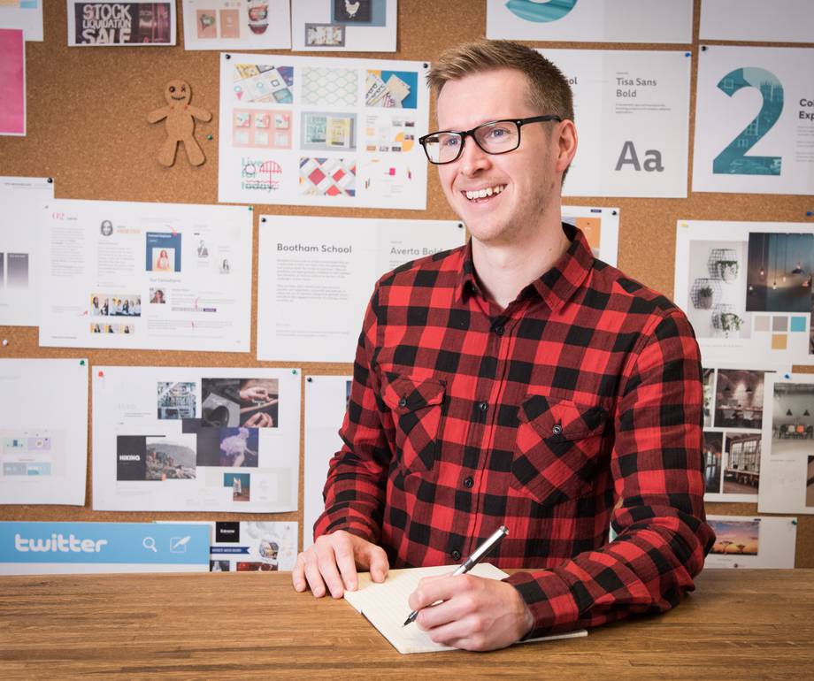 A smiling man with glasses in a red and black checkered shirt sitting at a desk with a notebook, in front of a bulletin board covered with design posters.
