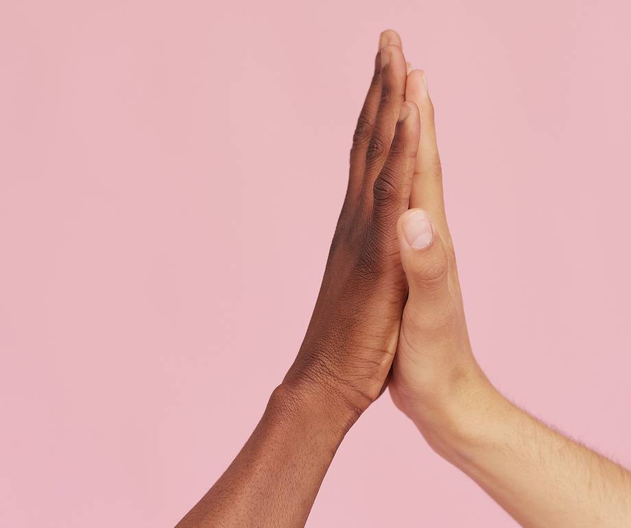 Two hands, one dark-skinned and one light-skinned, touching with palms pressed together against a pink background.