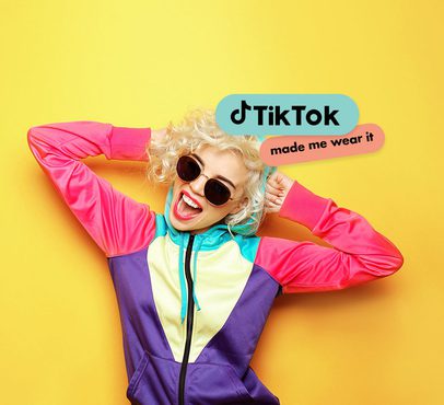 Woman with curly blonde hair, wearing colourful jacket and sunglasses, smiling against yellow background with TikTok and made me wear it stickers.