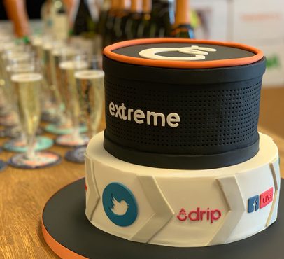Two-tiered cake designed as a portable speaker with social media icons, "extreme" branding, and a live streaming symbol, on a wooden table with glasses in the background.