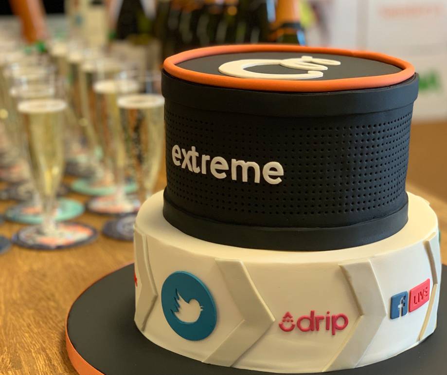 Two-tiered cake designed as a portable speaker with social media icons, "extreme" branding, and a live streaming symbol, on a wooden table with glasses in the background.