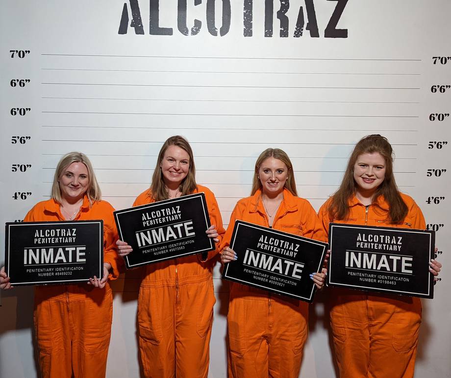 Four people in orange jumpsuits pose against a height chart backdrop marked "ALCOTRAZ", each holding a black "Alcotraz Penitentiary Inmate" sign.