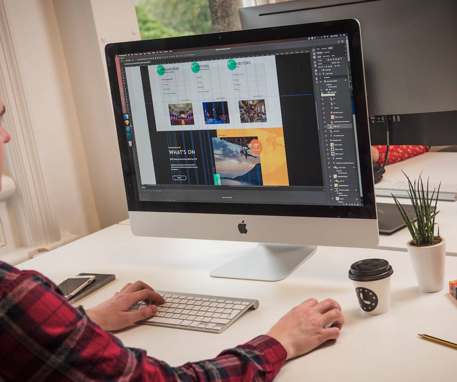 Person working on a website design on an iMac computer at a white desk with a coffee cup, plant, and other office items, in a room with natural light.