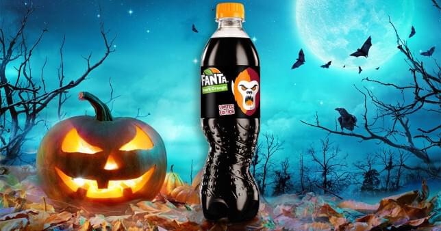 An image of a pumpkin next to a bottle of black fanta with a spooky background of a dark forrest.