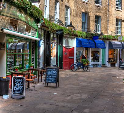 A pedestrian street with small shops and cafes, outdoor seating, colourful awnings, and potted plants, set against a brick building with windows and greenery.