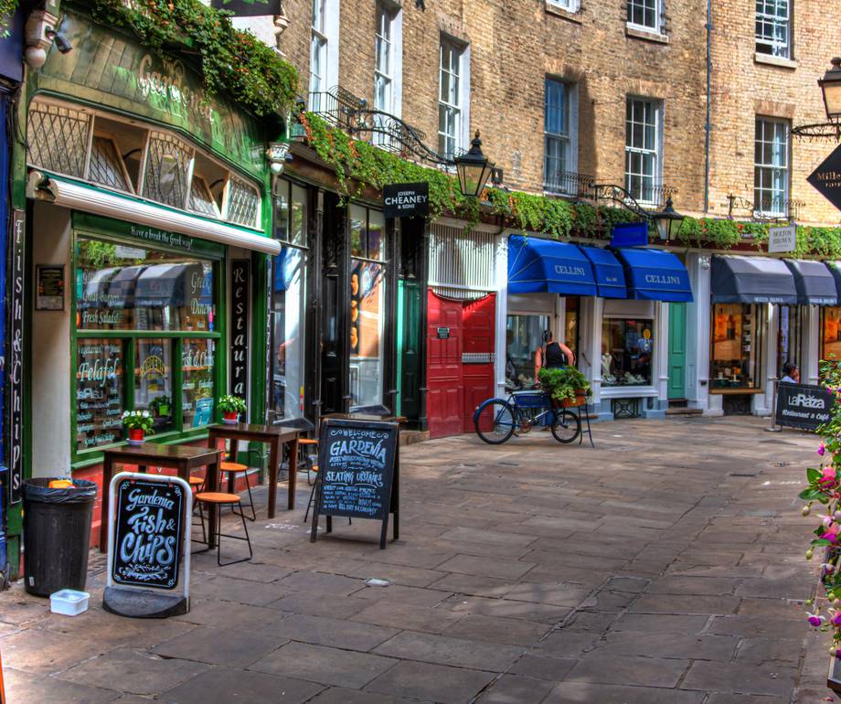 A pedestrian street with small shops and cafes, outdoor seating, colourful awnings, and potted plants, set against a brick building with windows and greenery.