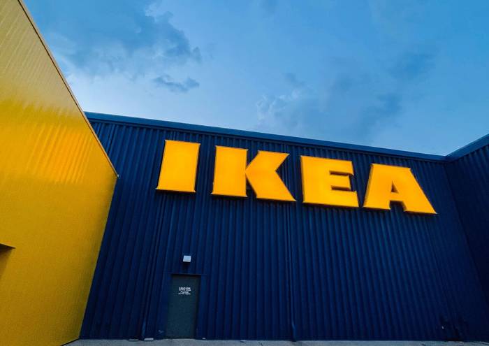 A low-angle view of an IKEA store exterior at dusk, with illuminated yellow IKEA letters on a dark blue corrugated wall beneath a blue sky, and a yellow wall to the left.