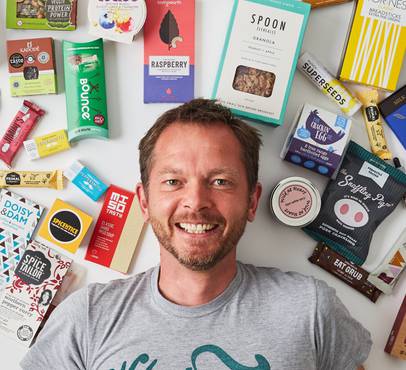 A smiling man lying on a white surface surrounded by various food and snack packages, with his head near the centre of the image.