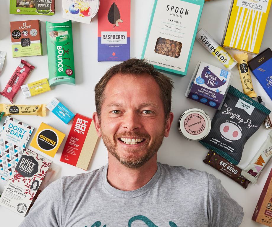 A smiling man lying on a white surface surrounded by various food and snack packages, with his head near the centre of the image.