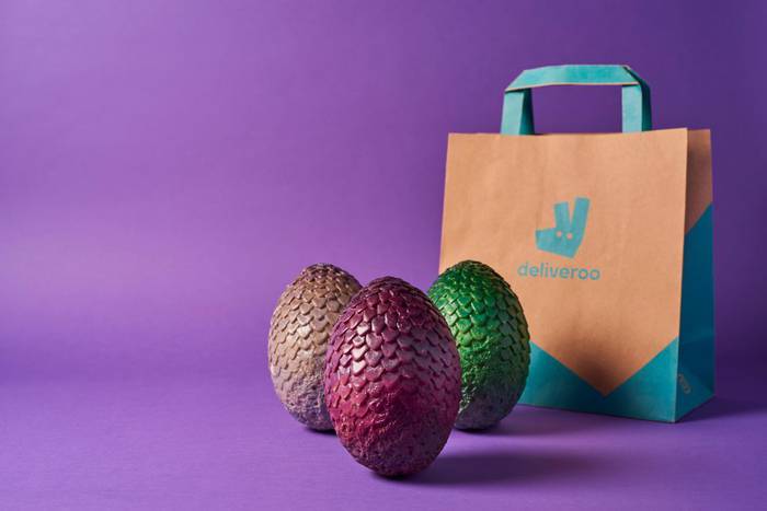 Three scaly dragon eggs in brown, magenta and green sit in front of a kraft paper Deliveroo bag with teal handles, against a plain violet backdrop
