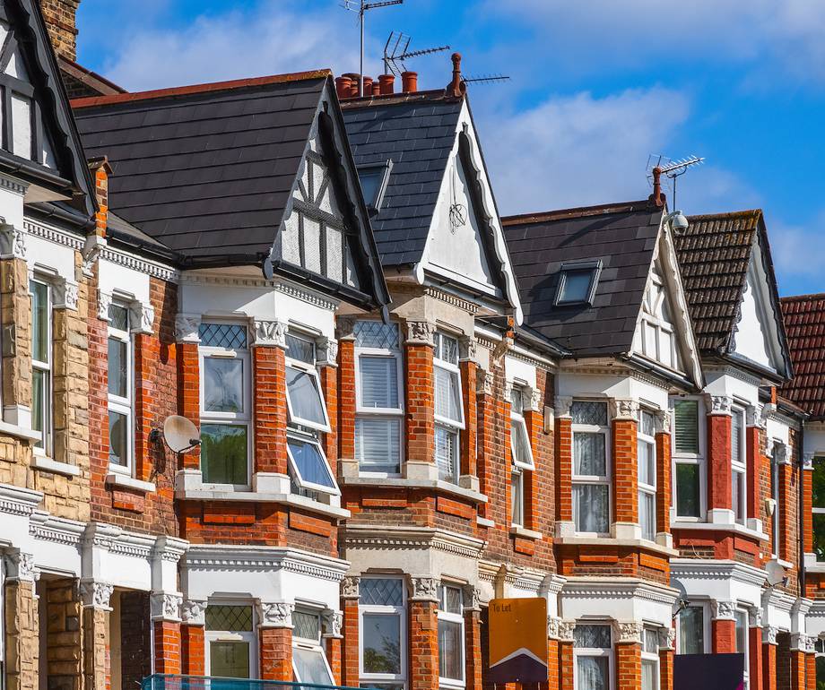 Row of ornate Victorian houses with red brick and white trim under a blue sky, showcasing varied rooftop designs and a "To Let" sign.