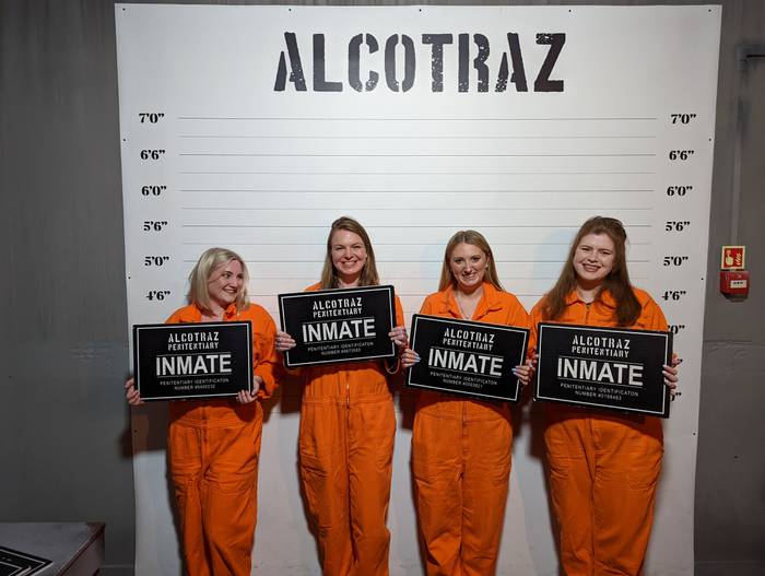 Four smiling people in orange jumpsuits hold black Alcotraz Penitentiary Inmate signs before a height chart backdrop, posing for a mugshot-style photo.