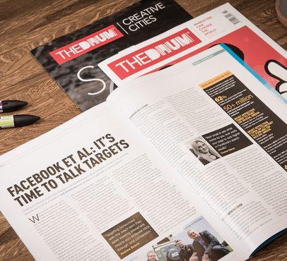 An open newspaper on a wooden table with an article titled "Facebook et al: It’s time to talk targets", surrounded by three pens and a magazine in the background.