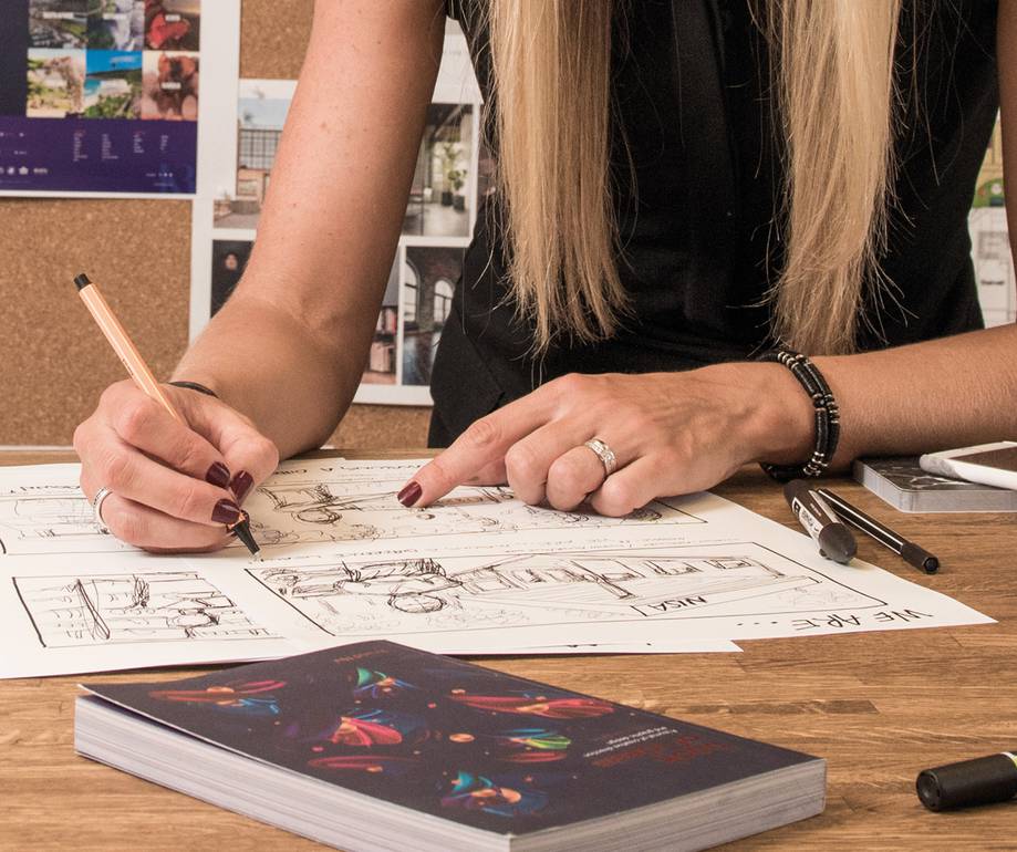 A woman with long blonde hair sketches on a large sheet of paper at a wooden table, with a book and a black marker nearby, in a creative workspace.
