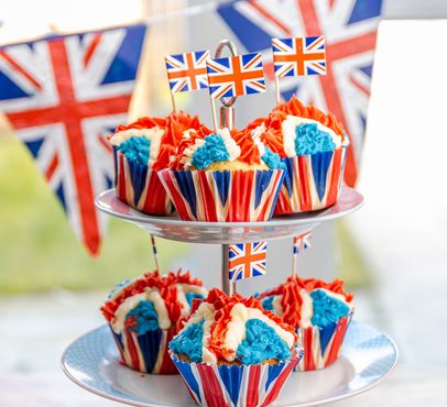 Cupcakes with red, white, and blue icing in Union Jack themed wrappers, displayed on a tiered stand with British flags in the background.