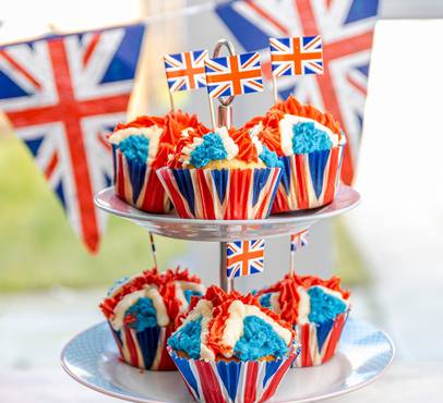 Cupcakes with red, white, and blue icing in Union Jack themed wrappers, displayed on a tiered stand with British flags in the background.