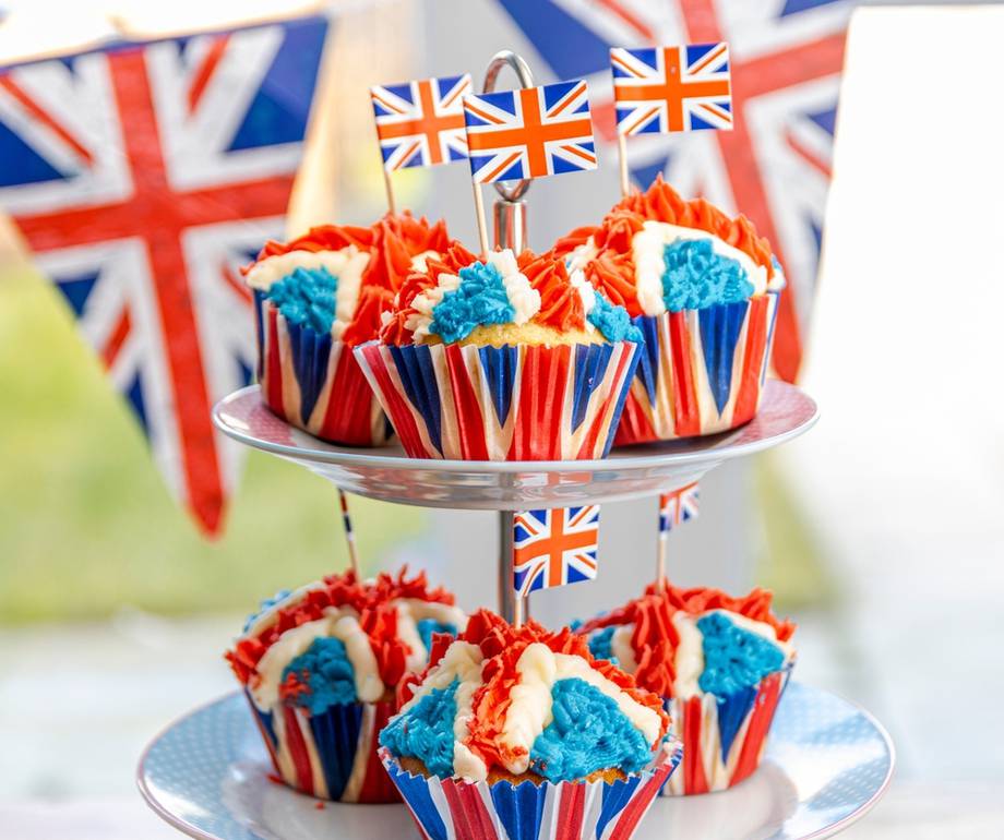 Cupcakes with red, white, and blue icing in Union Jack themed wrappers, displayed on a tiered stand with British flags in the background.