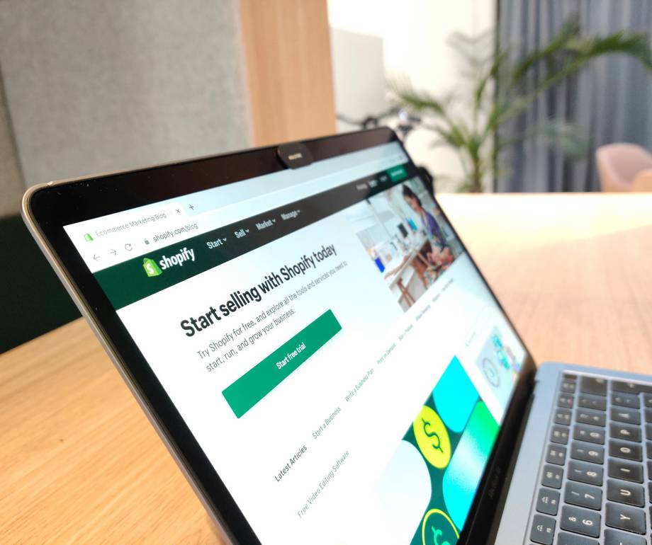 A close-up of a tablet displaying the Shopify homepage with a keyboard attached, on a wooden table in a bright office environment.