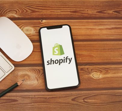 A smartphone displaying the Shopify logo on a wooden desk with a keyboard, mouse, pencil, and a small cactus plant.