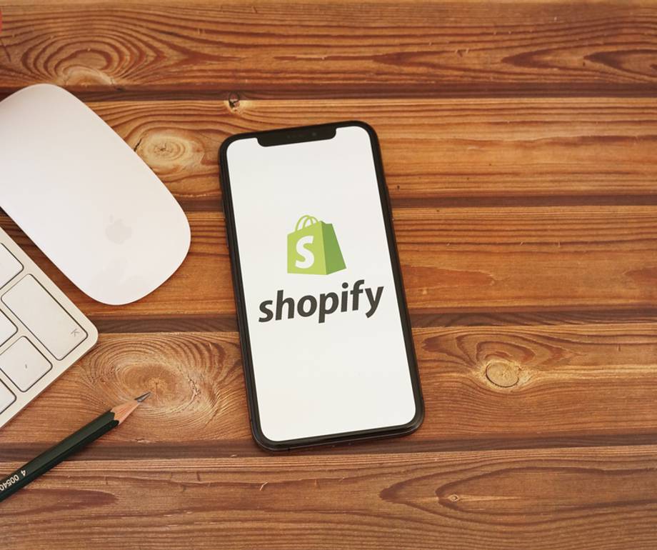 A smartphone displaying the Shopify logo on a wooden desk with a keyboard, mouse, pencil, and a small cactus plant.