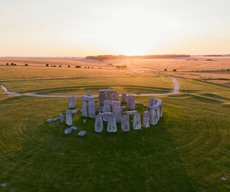 The image shows the ancient stone circle of Stonehenge at sunset, surrounded by green fields and winding paths, with a bright sky in the background.
