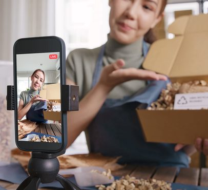 A woman holds a box of mixed nuts, recording a live stream on her smartphone, which is mounted on a tripod, in a bright indoor setting.