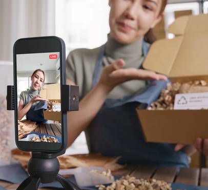 Smartphone on a tripod livestreaming a person in an apron showing an open cardboard box packed with recycled paper filling and a recycling label.