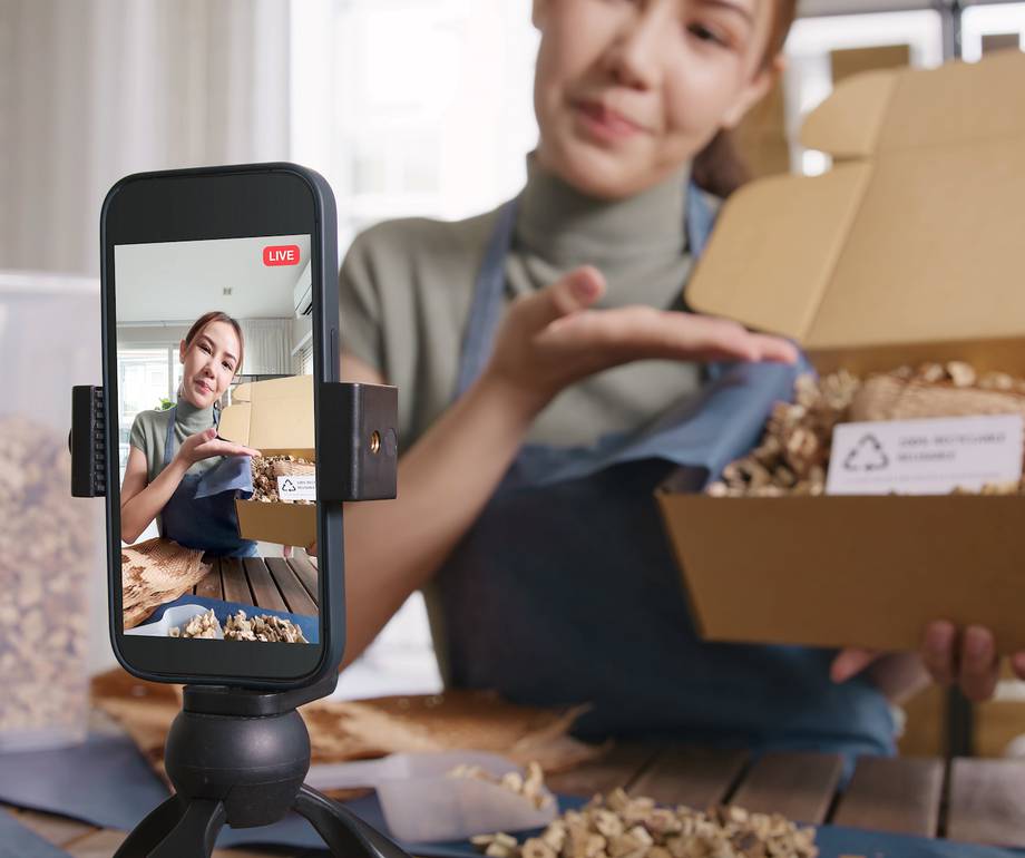 A woman holds a box of mixed nuts, recording a live stream on her smartphone, which is mounted on a tripod, in a bright indoor setting.