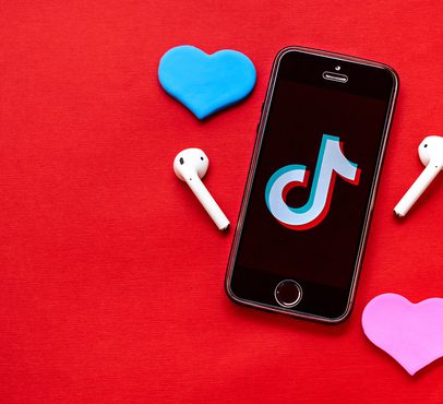 A smartphone displaying the TikTok logo surrounded by white wireless earbuds, with a blue heart-shaped object above and a pink heart-shaped object below on a red background.