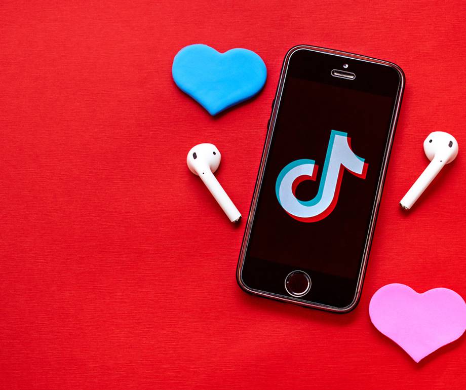 A smartphone displaying the TikTok logo surrounded by white wireless earbuds, with a blue heart-shaped object above and a pink heart-shaped object below on a red background.
