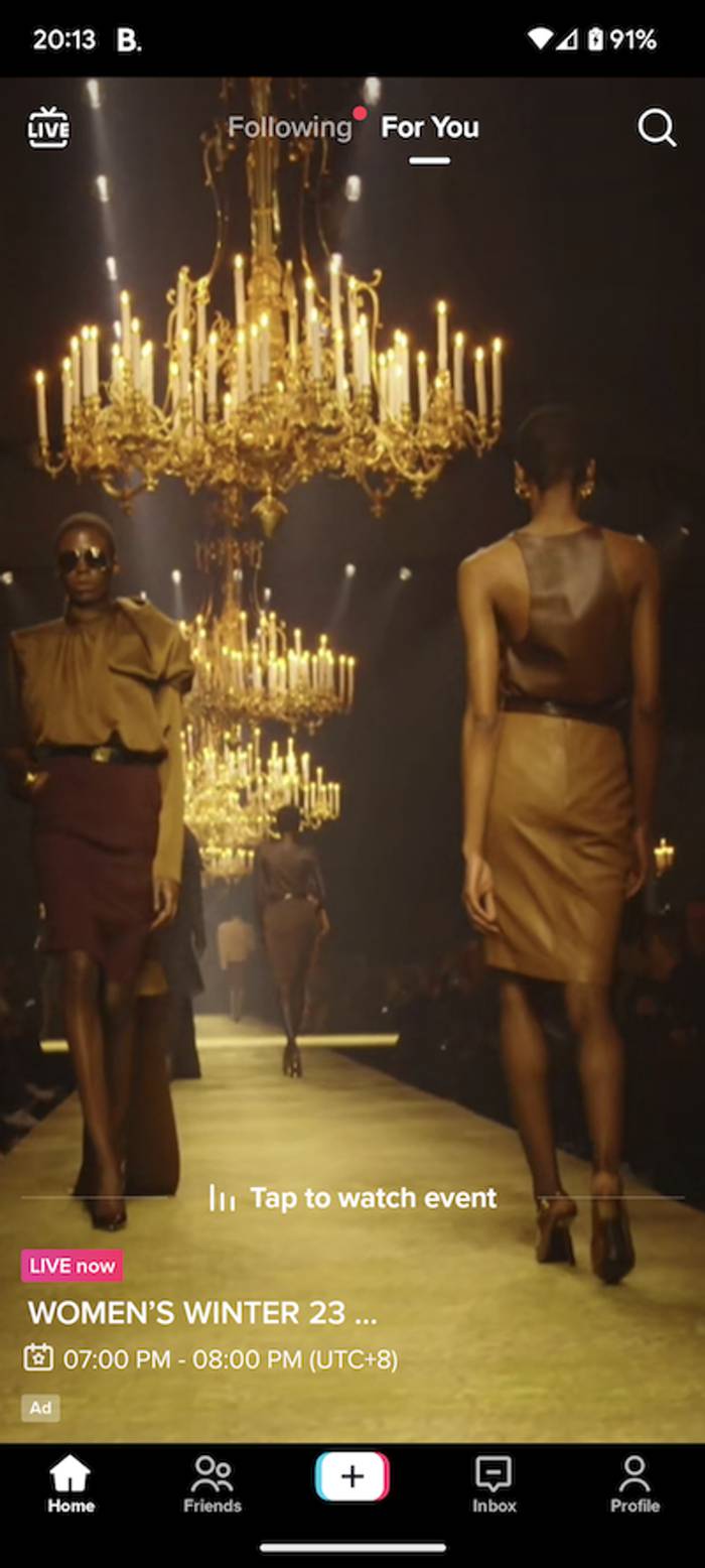 Phone screen showing a live fashion show: two models in tan and brown outfits walk past gold chandeliers on a golden runway, with event details overlaid.