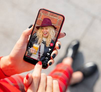 Hand with red nail polish holding a smartphone showing a smiling person with curly blonde hair, a burgundy hat and an orange drink, in front of a brick wall.