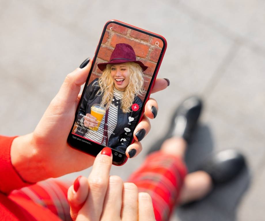 Person holding a phone capturing a woman with a hat holding a drink, smiling in front of a brick wall, with the person wearing red clothing and black shoes.