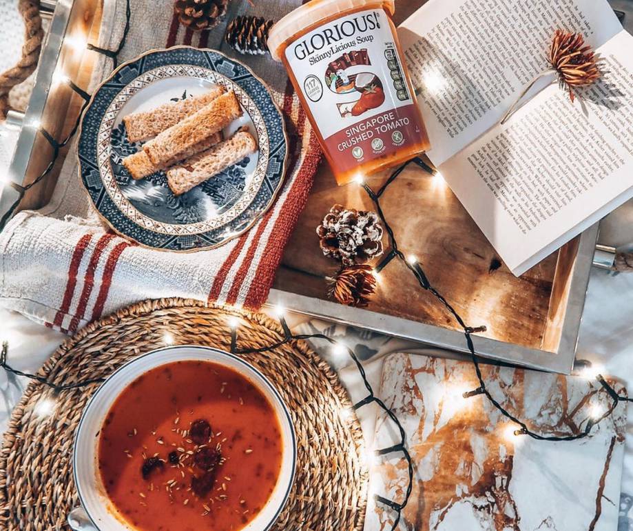 A top-down image of a cozy table setting with tomato soup, a plate of breadsticks, an open book, a jar of soup, decorative pinecones, and string lights.