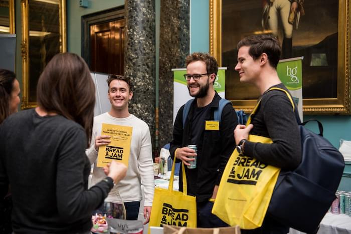 Four people talking at a conference holding yellow bags with the Bread and Jam logo on it.