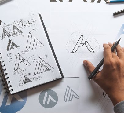 Person sketching a logo design on paper with sketches of logo concepts and geometric guides, surrounded by design notes and printed logo samples.