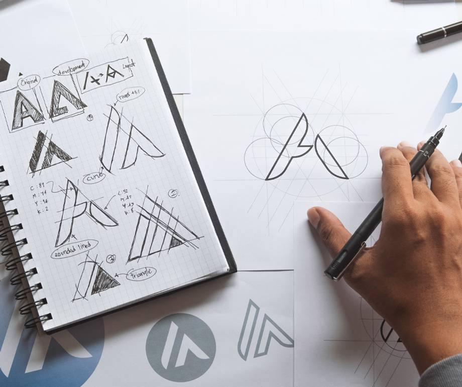Person sketching a logo design on paper with sketches of logo concepts and geometric guides, surrounded by design notes and printed logo samples.