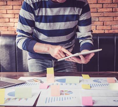 Person in striped sweater using a tablet at a cluttered table with charts, sticky notes, and a laptop against a brick wall.