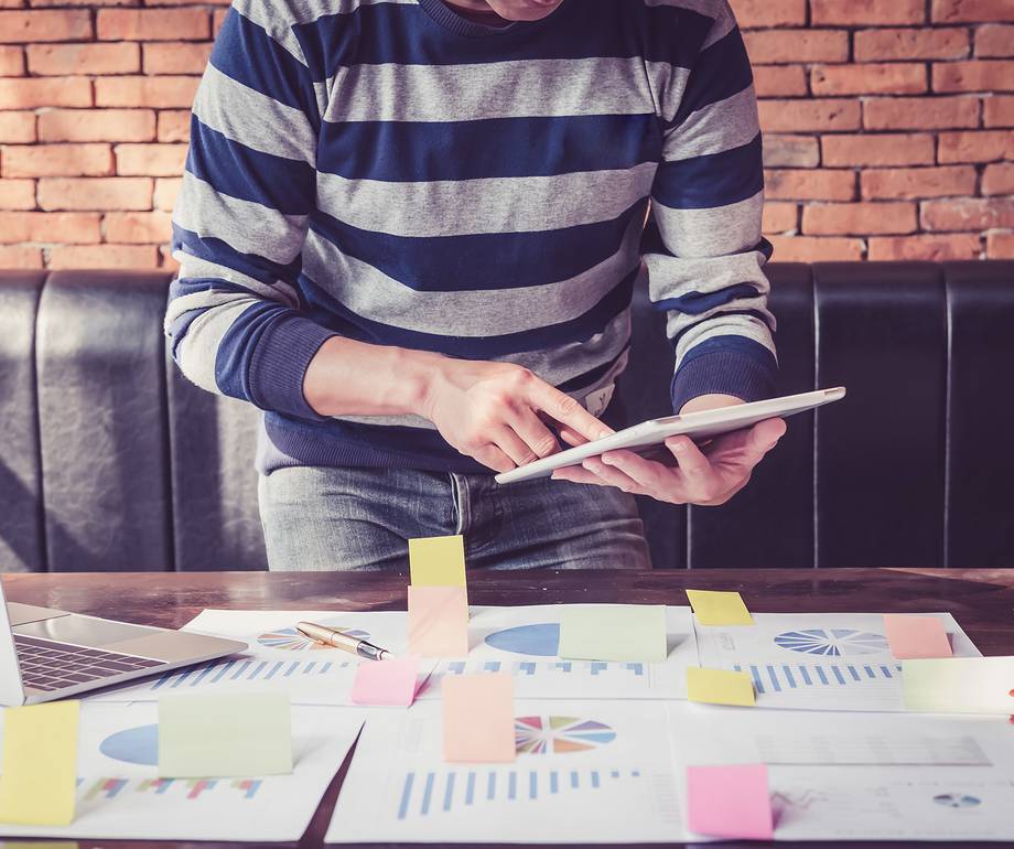 Person in striped sweater using a tablet at a cluttered table with charts, sticky notes, and a laptop against a brick wall.