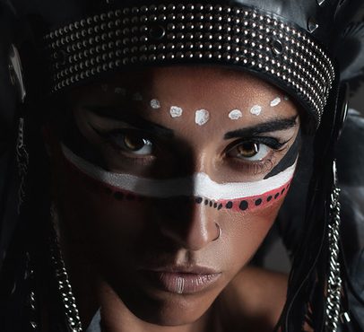 Close-up of a woman with intricate face paint, wearing a studded black headpiece, looking intensely at the camera with piercing eyes.
