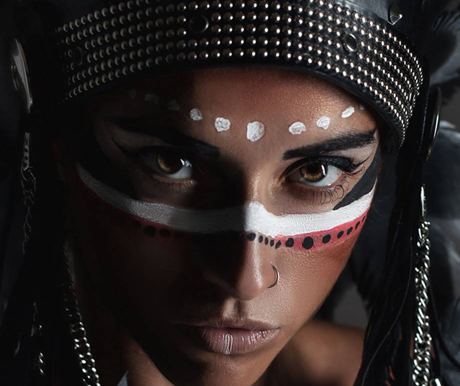 Close-up of a woman with intricate face paint, wearing a studded black headpiece, looking intensely at the camera with piercing eyes.