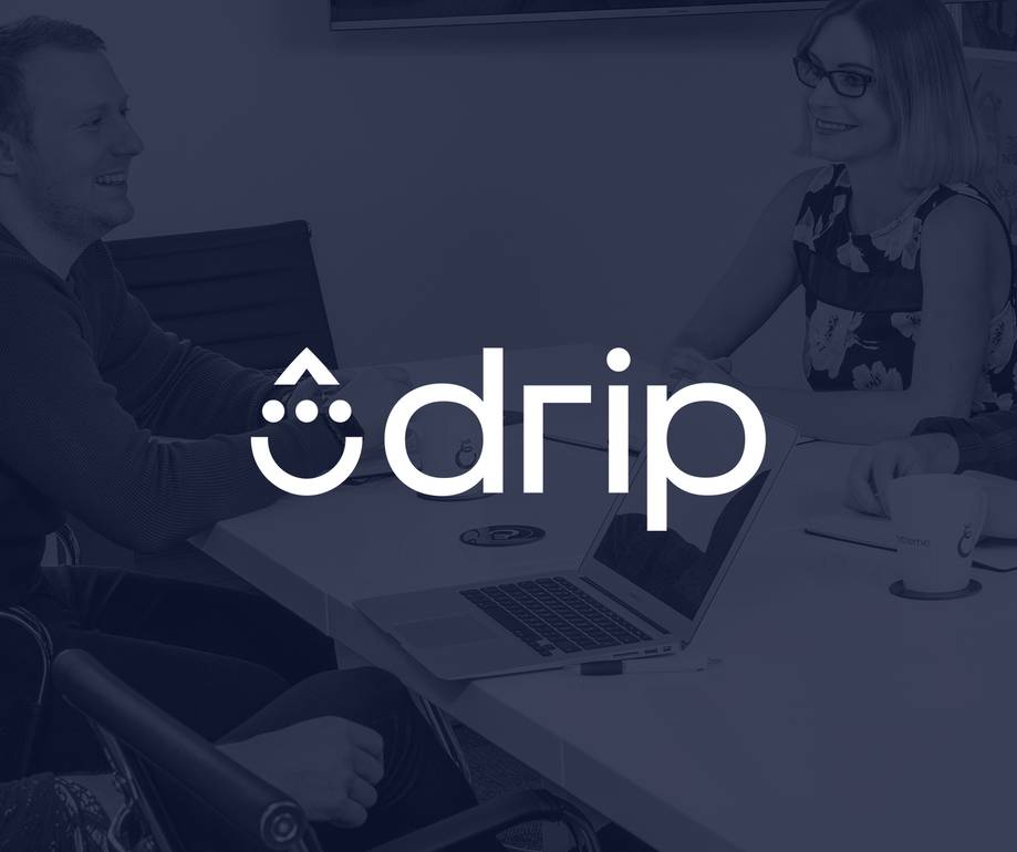 Two people smiling and conversing at a conference table with a laptop, coffee mugs, and a large screen in the background.