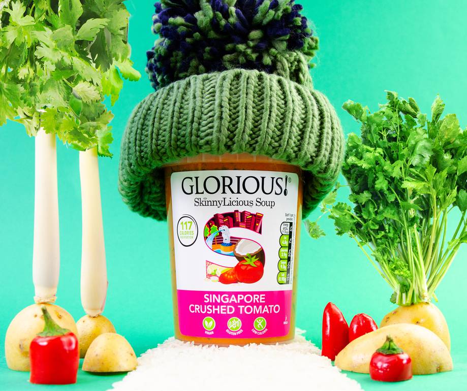 A jar of Glorious! Singapore Crushed Tomato soup wearing a green knitted beanie with a pom-pom, surrounded by vegetables against a turquoise background.