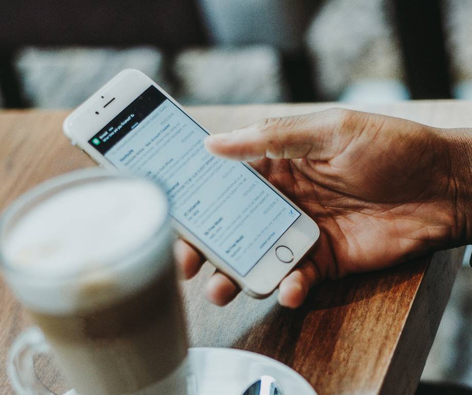 Hand holding a smartphone with a messaging app open, on a wooden table near a cup of coffee and a spoon in a cafe setting.