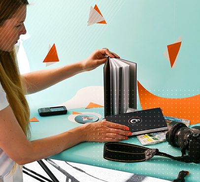 A woman with long hair is flipping through a photo album on a teal table, with a camera, remote, and colour swatches nearby, against a colourful background.