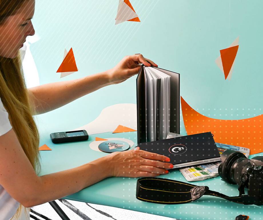 A woman with long hair is flipping through a photo album on a teal table, with a camera, remote, and colour swatches nearby, against a colourful background.