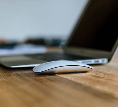 A white computer mouse in front of a laptop on a wooden surface, shot from a low angle with focus on the mouse.