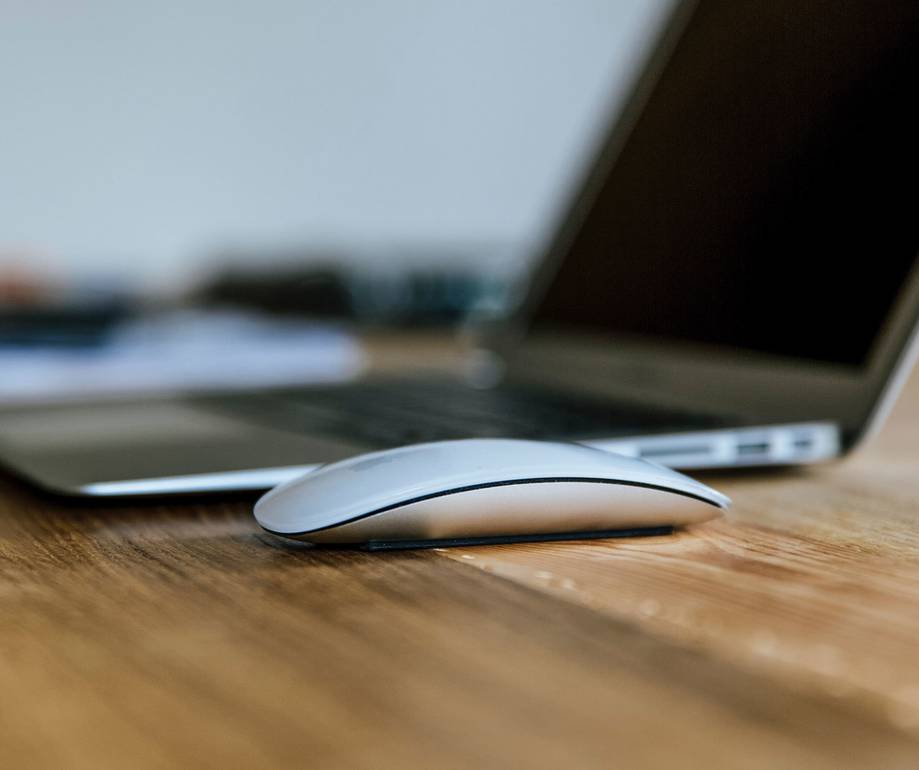 A white computer mouse in front of a laptop on a wooden surface, shot from a low angle with focus on the mouse.