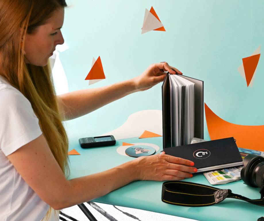 A woman with long hair arranging items on a blue table, including a black book, camera, and remote control, with geometric paper decorations in the background.