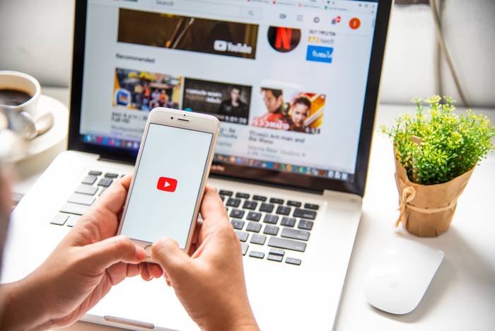 A picture of someone holding a mobile phone with the youtube logo on it, in front of a laptop which is displaying a list of video thumbnails on Youtube.